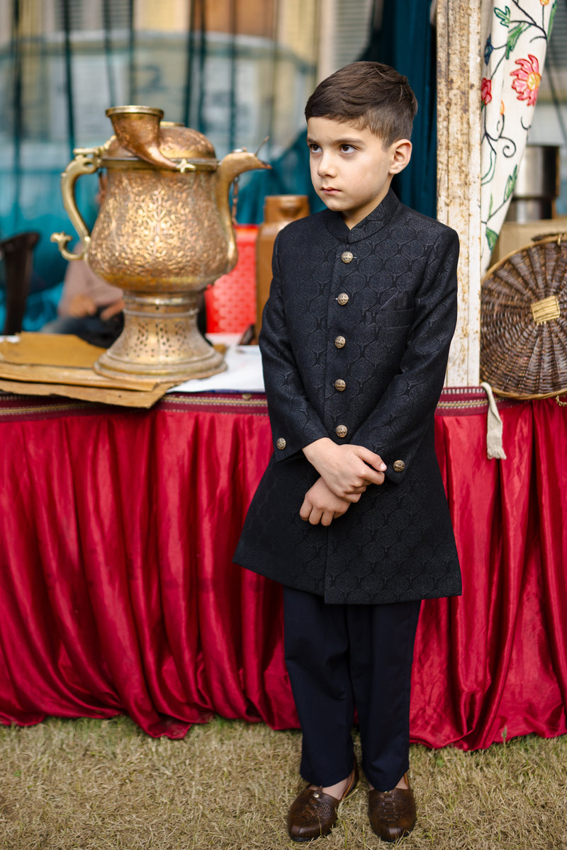 Young boy in formal attire standing next to a decorative urn with a red draped tablecloth in the background.